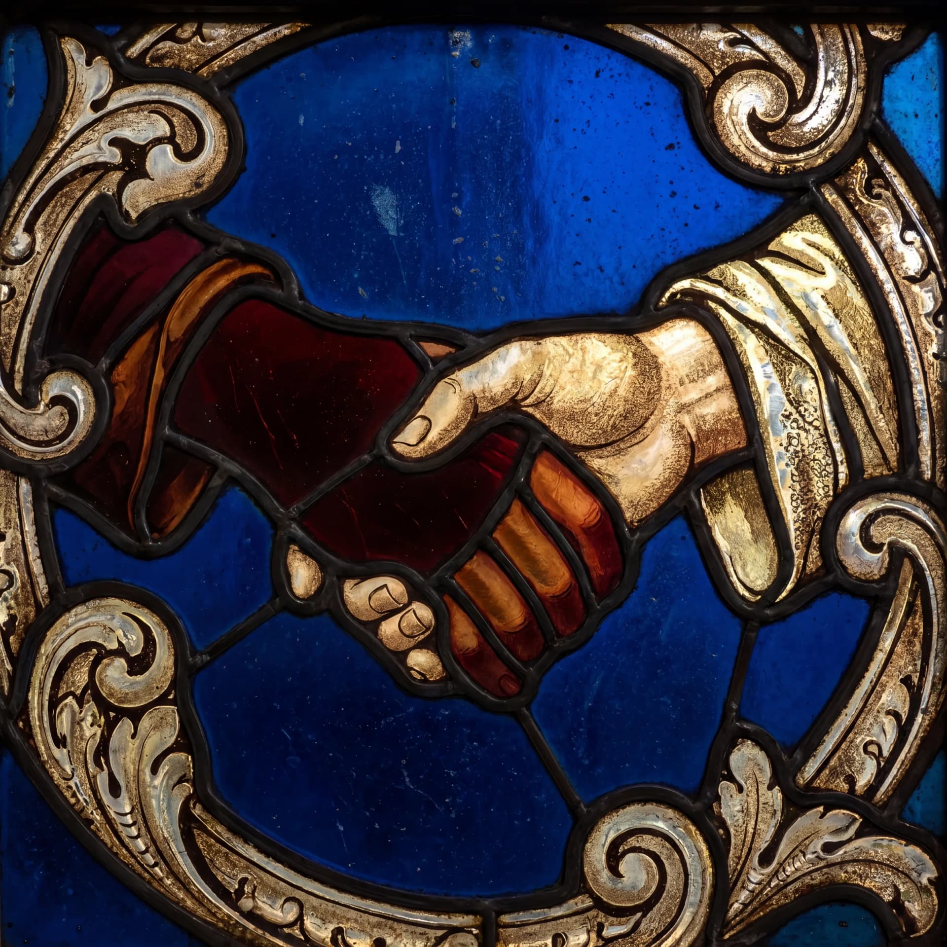 Stained glass panel of two clasped hands framed in gold scrollwork